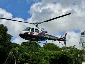 Fotografia em ângulo contraponto de um helicóptero da Polícia Militar do Estado de São Paulo (grupamento Águia) sobrevoando em baixa altitude. A aeronave possui pintura em branco, preto e detalhes em vermelho, com a inscrição "POLÍCIA MILITAR" em destaque na lateral do cauda. Ao fundo, vê-se um céu azul com nuvens brancas encorpadas, copas de árvores verdes logo abaixo do helicóptero e a lateral de um edifício urbano no canto direito.