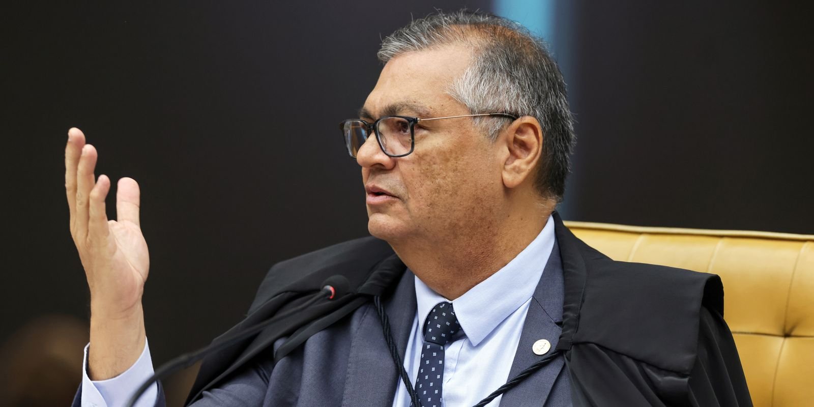 O ministro do Supremo Tribunal Federal (STF) Flávio Dino vestindo toga preta, terno cinza, camisa azul-clara e gravata escura, usando óculos de grau e gesticulando com a mão direita erguida enquanto fala.