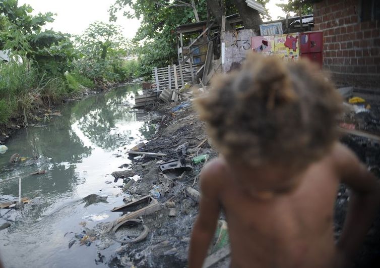 Imagem de riacho com falta de saneamento básico e uma criança passando em favela do Complexo da Maré