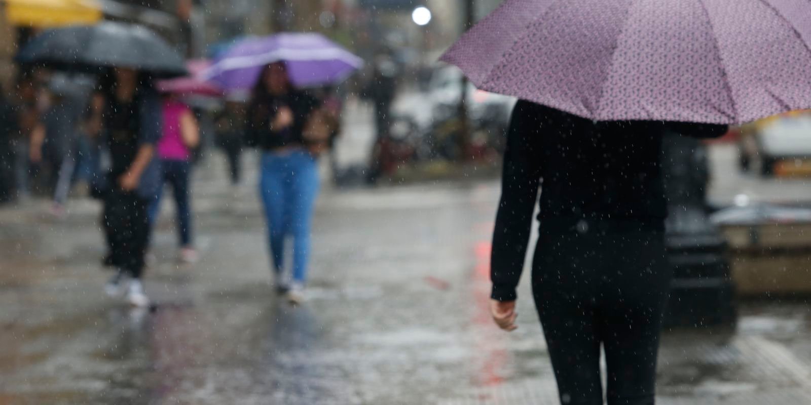 Fotografia que registra um momento de tempestade e chuva forte em uma via pública. O acumulado de água forma um grande ponto de alagamento na pista, refletindo a iluminação das proximidades e os pingos d'água sob o céu nublado e o tempo fechado.