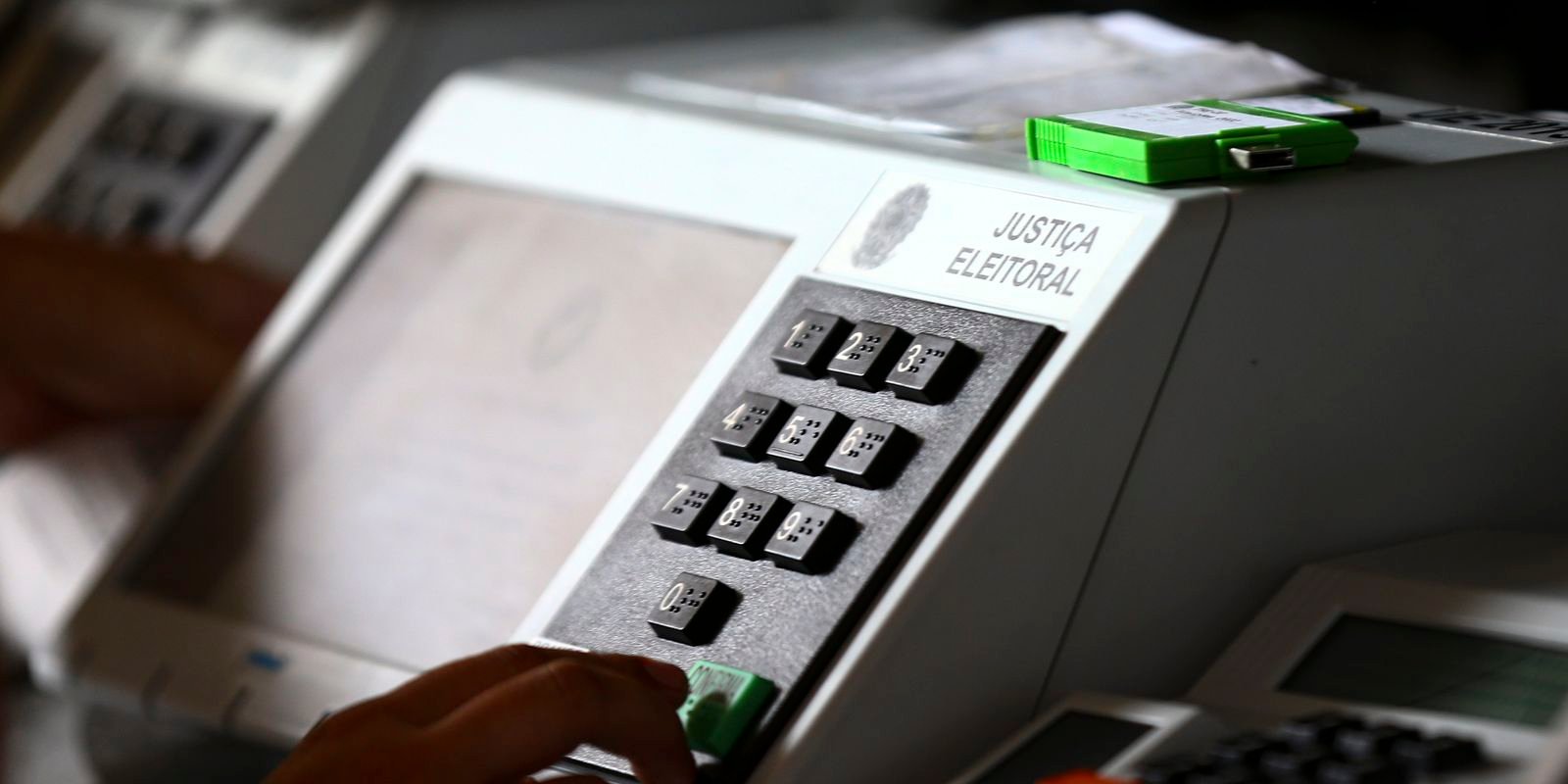 Urna eletrônica da Justiça Eleitoral brasileira em primeiro plano, com o teclado numérico e teclas coloridas visíveis. Uma pessoa digita um número na urna, enquanto outras urnas aparecem desfocadas ao fundo.