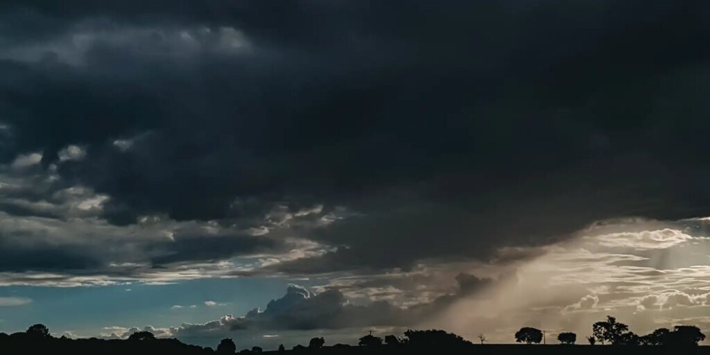 Fotografia de paisagem rural sob céu carregado com densas nuvens escuras de tempestade. No canto inferior direito, é possível observar raios de luz solar atravessando as nuvens e uma cortina de chuva caindo ao longe. Na linha do horizonte, destacam-se as silhuetas escuras de árvores e da vegetação contra o contraste da luz.