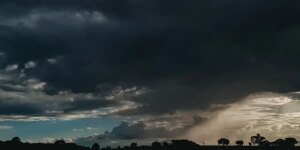 Fotografia de paisagem rural sob céu carregado com densas nuvens escuras de tempestade. No canto inferior direito, é possível observar raios de luz solar atravessando as nuvens e uma cortina de chuva caindo ao longe. Na linha do horizonte, destacam-se as silhuetas escuras de árvores e da vegetação contra o contraste da luz.