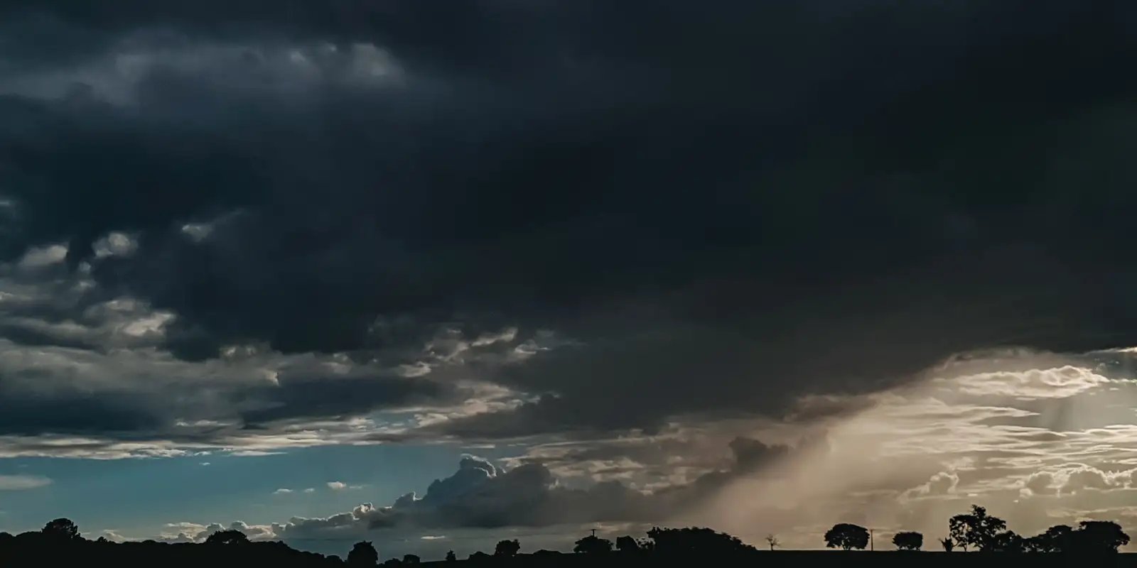 Fotografia de paisagem rural sob céu carregado com densas nuvens escuras de tempestade. No canto inferior direito, é possível observar raios de luz solar atravessando as nuvens e uma cortina de chuva caindo ao longe. Na linha do horizonte, destacam-se as silhuetas escuras de árvores e da vegetação contra o contraste da luz.