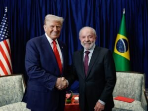 Fotografia oficial de encontro diplomático. À esquerda, Donald Trump, usando terno azul-marinho e gravata vermelha, sorri enquanto aperta a mão do presidente brasileiro Luiz Inácio Lula da Silva, posicionado à direita de terno escuro e gravata vinho. Ao fundo, sob cortinas azuis, estão dispostas a bandeira dos Estados Unidos (à esquerda) e a bandeira do Brasil (à direita).