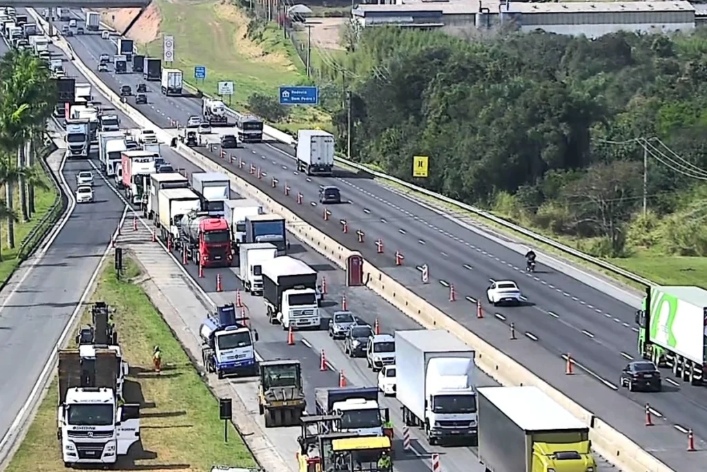 Vista aérea de trecho da Rodovia Dom Pedro I em dia ensolarado. No sentido esquerdo, há retenção de tráfego e filas de caminhões e carros dividindo espaço com cones de sinalização e maquinário de obras asfálticas. No sentido oposto, à direita, o trânsito flui livremente com poucos veículos. A rodovia é cercada por vegetação e áreas verdes.