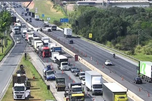 Vista aérea de trecho da Rodovia Dom Pedro I em dia ensolarado. No sentido esquerdo, há retenção de tráfego e filas de caminhões e carros dividindo espaço com cones de sinalização e maquinário de obras asfálticas. No sentido oposto, à direita, o trânsito flui livremente com poucos veículos. A rodovia é cercada por vegetação e áreas verdes.