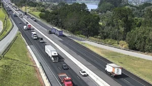 Vista aérea da Rodovia D. Pedro I em um trecho de pista dupla, com três faixas de cada lado, com tráfego moderado de veículos, incluindo caminhões de carga e carros de passeio. As pistas são separadas por uma barreira de concreto, e a rodovia é cercada por gramados verdes, áreas arborizadas e uma represa visível ao fundo, sob céu limpo.
