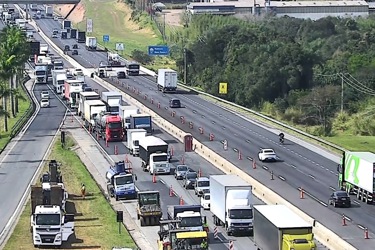 Vista aérea de trecho da Rodovia Dom Pedro I em dia ensolarado. No sentido esquerdo, há retenção de tráfego e filas de caminhões e carros dividindo espaço com cones de sinalização e maquinário de obras asfálticas. No sentido oposto, à direita, o trânsito flui livremente com poucos veículos. A rodovia é cercada por vegetação e áreas verdes.