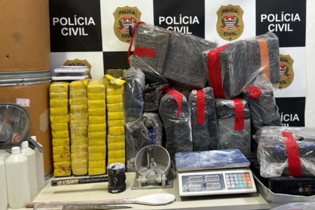 Diversos tabletes de drogas apreendidos dispostos sobre uma mesa, incluindo pilhas de tabletes embalados em plástico amarelo e outros maiores envolvidos em plástico transparente e fita vermelha ou preta. Ao redor dos entorpecentes, há insumos e utensílios como balança de precisão, faca, colher grande com resíduos de pó, frascos de produtos químicos, martelo e uma prensa industrial. Ao fundo, destaca-se um painel xadrez em preto e branco com o brasão e a inscrição "Polícia Civil.