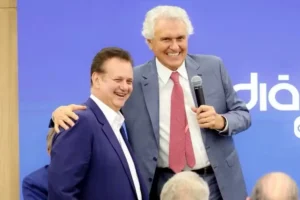 Gilberto Kassab, vestindo terno azul-marinho e camisa branca sem gravata, sorri ao lado do governador Ronaldo Caiado, que veste terno cinza, camisa branca e gravata vermelha. Caiado abraça Kassab pelos ombros e segura um microfone com a mão esquerda enquanto sorri. Ao fundo, há um painel azul com letras brancas.