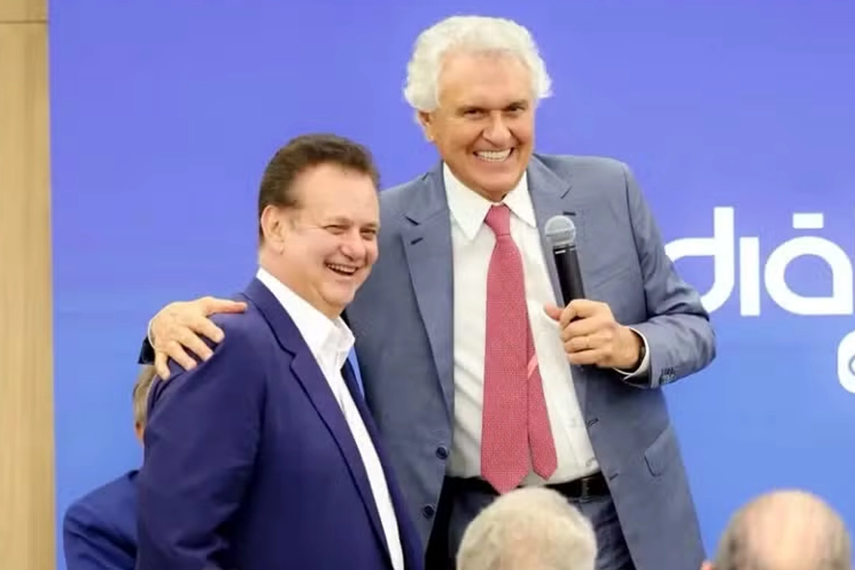 Gilberto Kassab, vestindo terno azul-marinho e camisa branca sem gravata, sorri ao lado do governador Ronaldo Caiado, que veste terno cinza, camisa branca e gravata vermelha. Caiado abraça Kassab pelos ombros e segura um microfone com a mão esquerda enquanto sorri. Ao fundo, há um painel azul com letras brancas.