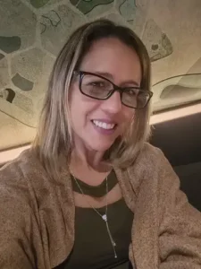 Selfie em ambiente interno de uma mulher de pele clara, sorrindo para a câmera. Ela usa óculos de grau com armação retangular escura, tem cabelos loiros de tom médio na altura dos ombros e veste uma blusa verde-oliva com decote vazado, combinada com um xale ou casaco bege sobreposto. Ela também usa um colar fino prateado com um pingente de pedra brilhante. Ao fundo, há uma parede decorada com textura em tons neutros de bege e verde.