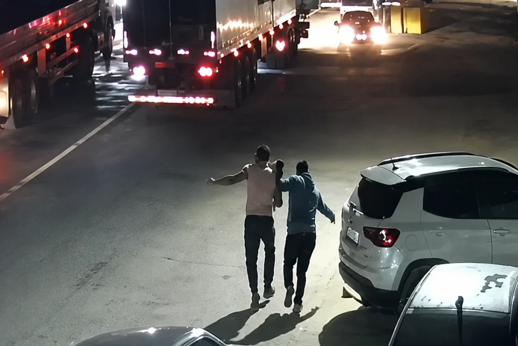Imagem de câmera de segurança mostra, à noite, dois homens caminhando lado a lado em uma rua movimentada, entre caminhões e carros estacionados. Um deles parece apoiar ou conduzir o outro pelo braço. A via está iluminada por faróis e lanternas de veículos.