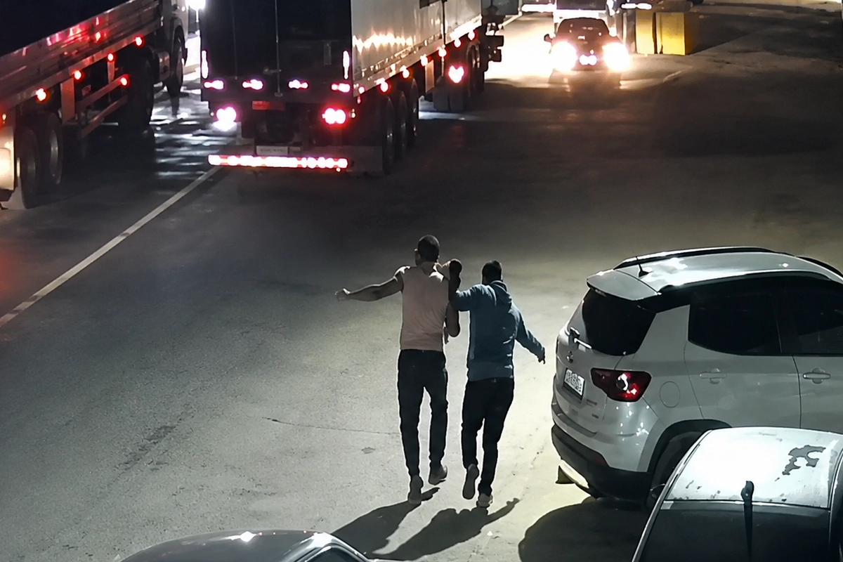 Imagem de câmera de segurança mostra, à noite, dois homens caminhando lado a lado em uma rua movimentada, entre caminhões e carros estacionados. Um deles parece apoiar ou conduzir o outro pelo braço. A via está iluminada por faróis e lanternas de veículos.
