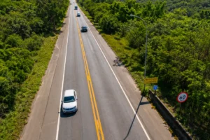 Rodovia Edgard Máximo Zambotto, em um trecho de duas pistas cercada por uma área de mata verde e montanhas ao fundo, vista de um ângulo aéreo. Vários veículos trafegam pela estrada, que possui faixa dupla amarela no centro e sinalização de limite de velocidade de 60 km/h. À direita, há uma placa de fiscalização eletrônica, um poste de iluminação e uma barreira de concreto.