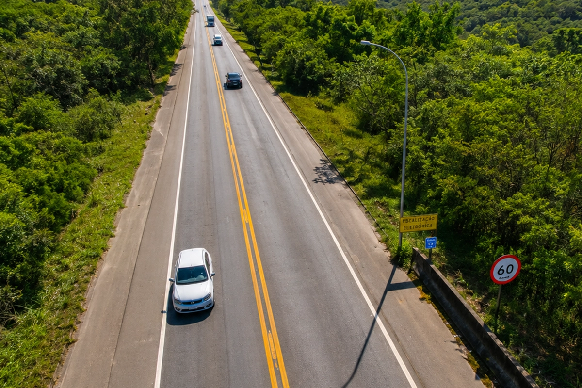 Rodovia Edgard Máximo Zambotto, em um trecho de duas pistas cercada por uma área de mata verde e montanhas ao fundo, vista de um ângulo aéreo. Vários veículos trafegam pela estrada, que possui faixa dupla amarela no centro e sinalização de limite de velocidade de 60 km/h. À direita, há uma placa de fiscalização eletrônica, um poste de iluminação e uma barreira de concreto.