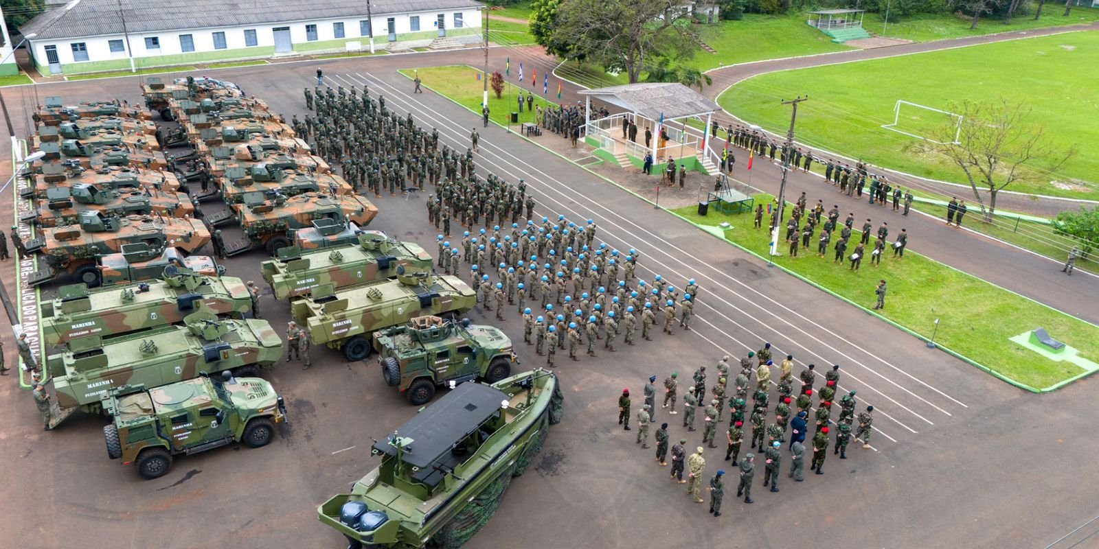 Fotografia em ângulo elevado (vista aérea) mostrando uma formação militar ao ar livre. À esquerda, há dezenas de veículos blindados e táticos camuflados estacionados em fileiras, incluindo uma embarcação militar em primeiro plano. No centro e à direita, centenas de militares em fardamento operacional e boinas (incluindo capacetes azuis de missão de paz) estão perfilados em formação rígida no pátio pavimentado. Ao fundo, veem-se arquibancadas, um campo gramado e instalações militares sob luz natural.