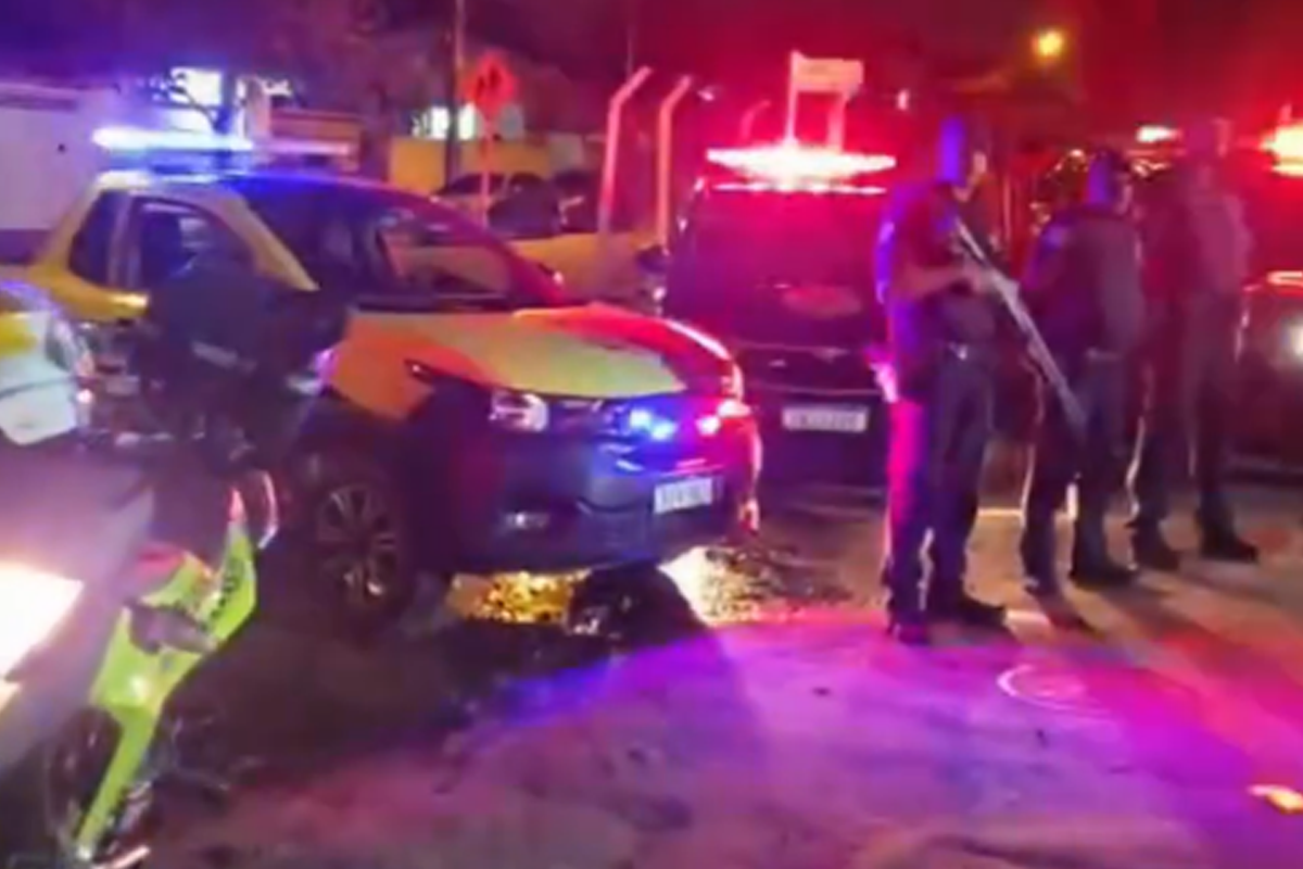 Policiais em pé à noite ao lado de viaturas e motocicletas com luzes de emergência azuis e vermelhas acesas.