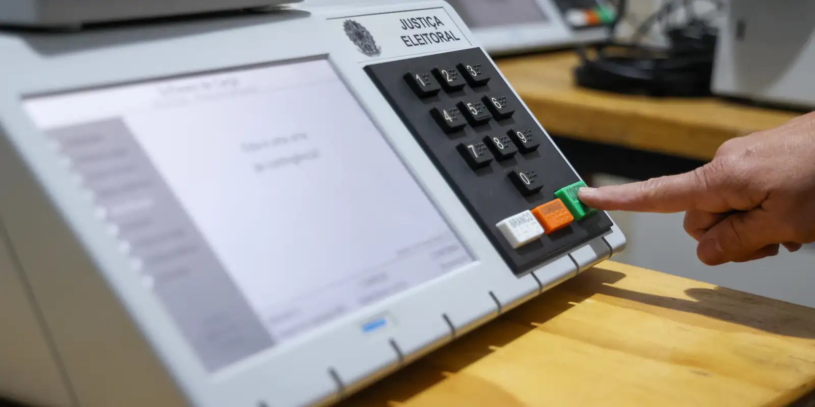 Foto em plano próximo de uma urna eletrônica brasileira sobre uma mesa de madeira. A tela exibe as opções de voto desfocadas. No topo do teclado numérico preto está gravado o brasão da República e o texto "JUSTIÇA ELEITORAL". Na parte inferior do teclado, o dedo indicador de um eleitor é visto confirmando o voto ao apertar o botão verde "CONFIRMA", posicionado ao lado dos botões "BRANCO" (branco) e "CORRIGE" (laranja).