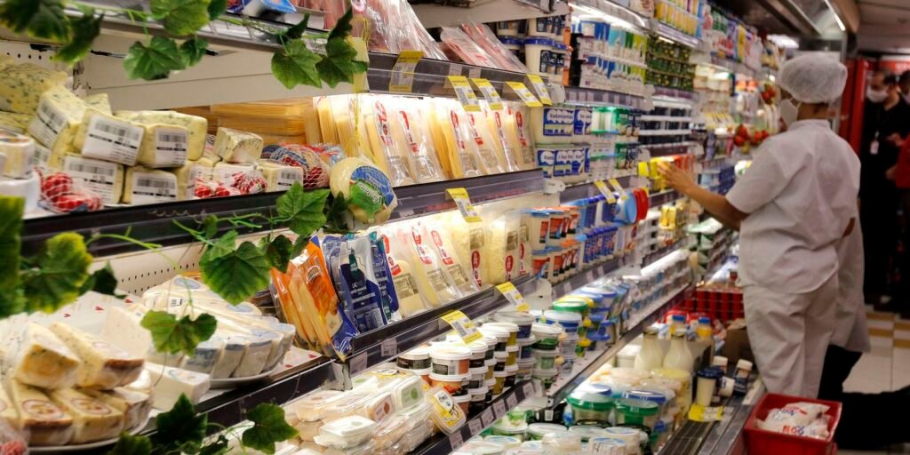 Vista de um corredor de supermercado focado na seção de laticínios e frios. As prateleiras e balcões refrigerados estão repletos de queijos fatiados, peças em embalagens plásticas e potes de iogurte ou requeijão. À direita, uma funcionária vestindo uniforme branco, touca protetora e máscara de proteção observa e organiza os produtos na prateleira. Folhagens artificiais decoram a borda dos expositores de vidro.