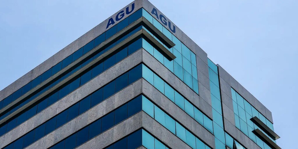 Fachada moderna de um edifício público de concreto e vidros espelhados azuis, vista de baixo para cima contra o céu limpo, exibindo a sigla AGU (Advocacia-Geral da União) no topo das duas faces do prédio.