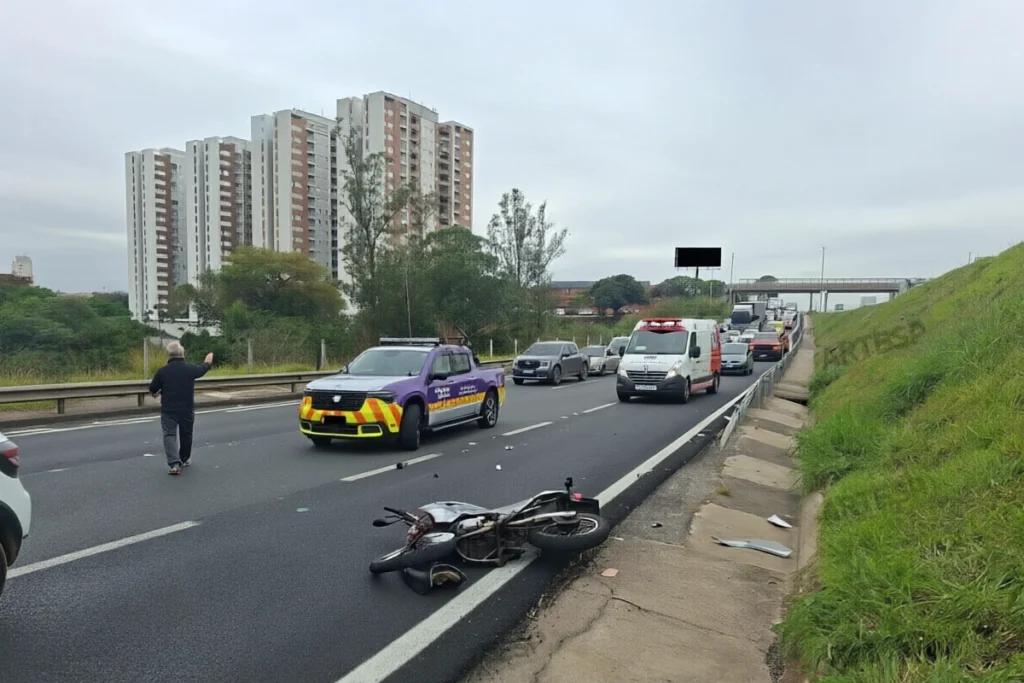 Cena de acidente de trânsito na rodovia Anhaguera perto de prédios residenciais, com uma motocicleta caída na pista, veículos de resgate e concessionária parados ao fundo e um trânsito congestionado ao longe.