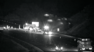 Imagem noturna em preto e branco capturada por câmera de segurança viária em uma rodovia. Vários veículos de resgate e carros estão parados no acostamento e parte da pista com luzes acesas e sinalização de emergência. O tráfego segue em velocidade reduzida ao lado da área bloqueada, sob iluminação dos faróis dos veículos.