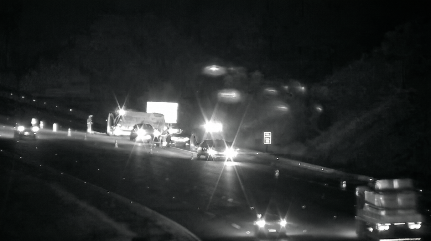Imagem noturna em preto e branco capturada por câmera de segurança viária em uma rodovia. Vários veículos de resgate e carros estão parados no acostamento e parte da pista com luzes acesas e sinalização de emergência. O tráfego segue em velocidade reduzida ao lado da área bloqueada, sob iluminação dos faróis dos veículos.