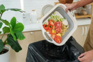 Pessoa usando avental marrom descartando restos de alimentos frescos, como fatias de tomate, pepino e cascas, de um recipiente retrátil diretamente em um saco de lixo preto numa cozinha.