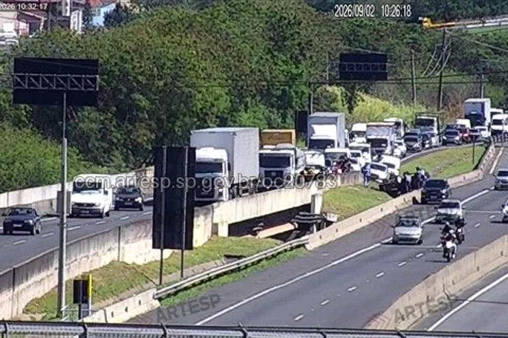 Imagem de uma câmera de monitoramento viário da ARTESP em uma rodovia. O tráfego no sentido esquerdo/superior está bastante congestionado, com uma longa fila de caminhões, vans e carros parados ou em ritmo lento. Na pista oposta, no lado direito, o trânsito flui normalmente com poucos veículos em movimento. No canteiro central entre as pistas, há um viaduto/passagem inferior e veículos estacionados. O dia está ensolarado e há uma vegetação densa ao fundo.