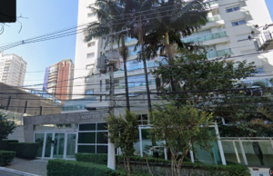 Fachada de um edifício residencial moderno em tom claro, com a identificação "L'ADRESSE 55" na portaria de entrada. Em frente ao prédio, há palmeiras, vegetação ornamental e um poste com fiação elétrica e transformador.