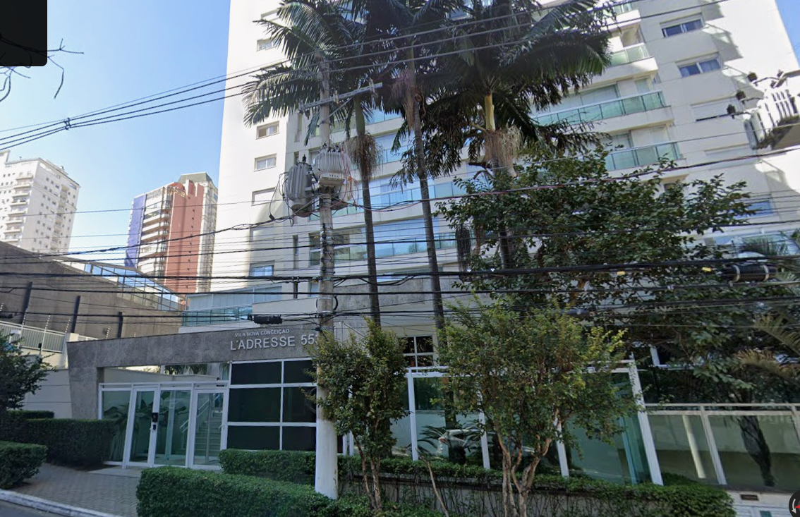 Fachada de um edifício residencial moderno em tom claro, com a identificação "L'ADRESSE 55" na portaria de entrada. Em frente ao prédio, há palmeiras, vegetação ornamental e um poste com fiação elétrica e transformador.