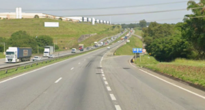 Fotografia panorâmica de uma rodovia asfaltada com faixas bem sinalizadas em um dia ensolarado. Na pista contrária, observa-se tráfego moderado com caminhões de carga e carros de passeio. Ao fundo e nas laterais, veem-se galpões industriais no topo de um barranco gramado, placas azuis de sinalização viária e vegetação nativa à beira da via.