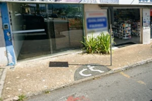 Fachada de estabelecimento comercial e farmácia vista da calçada, com destaque para a sinalização de acessibilidade desenhada no chão com o símbolo de cadeirante. Ao lado, há uma placa de trânsito fincada e plantas ornamentais em frente à vitrine.
