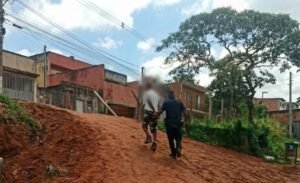 Fotografia que mostra um guarda municipal fardado de costas, conduzindo um homem algemado por uma ladeira de terra vermelha. O homem detido está com o rosto desfocado para preservação de identidade. Ao fundo, veem-se casas residenciais, fiação elétrica e árvores em um dia ensolarado com nuvens no céu.