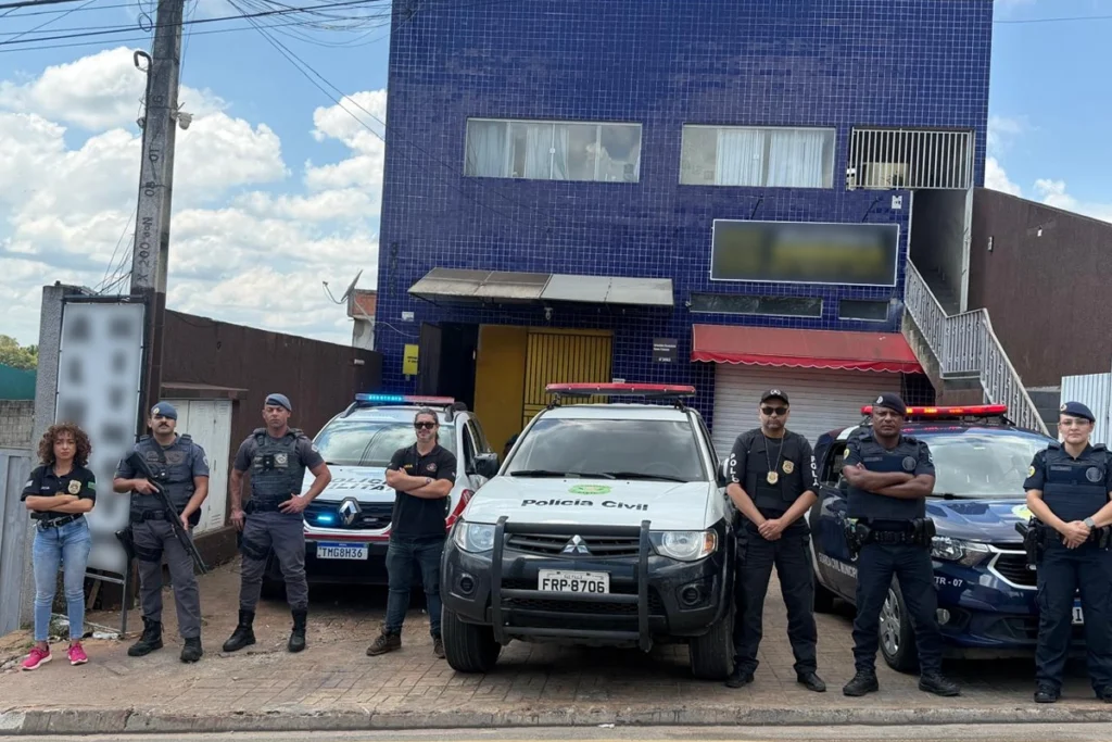 Fotografia de operação conjunta com policiais civis, policiais militares e guardas civis municipais em pé, de braços cruzados ou em posição de prontidão, alinhados em frente a três viaturas estacionadas. Ao centro está uma caminhonete branca da Polícia Civil, acompanhada por viaturas da Polícia Militar e da Guarda Municipal. Ao fundo, destaca-se o prédio da delegacia com fachada revestida de pastilhas azuis sob céu ensolarado.