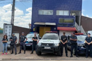 Fotografia de operação conjunta com policiais civis, policiais militares e guardas civis municipais em pé, de braços cruzados ou em posição de prontidão, alinhados em frente a três viaturas estacionadas. Ao centro está uma caminhonete branca da Polícia Civil, acompanhada por viaturas da Polícia Militar e da Guarda Municipal. Ao fundo, destaca-se o prédio da delegacia com fachada revestida de pastilhas azuis sob céu ensolarado.