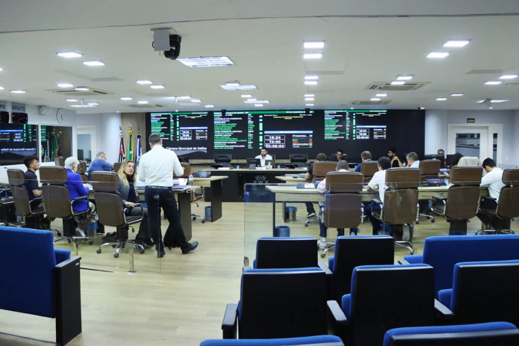 Vista ampla do plenário da Câmara Municipal de Itatiba a partir da galeria de auditório, em primeiro plano com poltronas azuis. Ao fundo, vereadores e assessores estão dispostos em bancadas de madeira com cadeiras de couro executivas em formato de "U". Um homem de camisa branca e calça escura está em pé no centro do plenário conversando com uma vereadora sentada. A parede de fundo exibe grandes painéis eletrônicos com telas pretas contendo a lista dos vereadores presentes, votações, horários e cronômetros da sessão.