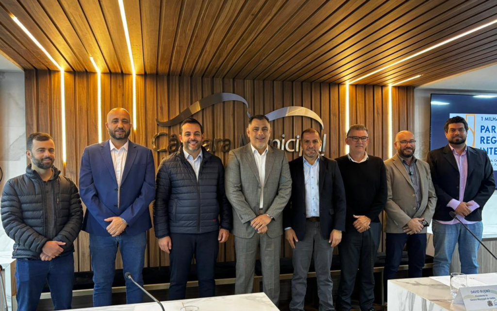 Oito homens vestindo trajes esporte fino e terno estão posados lado a lado em pé (vereador de Jundiaí Juninho Adilson, presidente da Câmara de Jarinu Rogério Sapão, prefeito de Jundiaí Gustavo Martinelli, presidente da Câmara de Itatiba Davi Bueno, presidente da Câmara de Jundiaí Edicarlos Vieira, presidente da Câmara de Várzea Paulista Dr. Elizeu Notário, presidente da Câmara de Louveira Kaká Rodrigues e o vereador de Jundiaí Tiago Leandro). Eles se encontram no plenário da Câmara Municipal de Jundiaí, em frente a um painel de madeira com fita de LED embutida e a inscrição do logotipo da instituição ao fundo. À direita, há uma tela exibindo uma apresentação. Na parte inferior em primeiro plano, vê-se a borda de uma bancada clara de mármore com microfones.