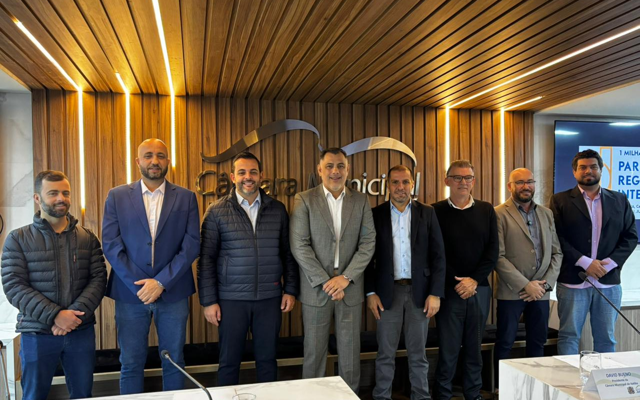 Oito homens vestindo trajes esporte fino e terno estão posados lado a lado em pé (vereador de Jundiaí Juninho Adilson, presidente da Câmara de Jarinu Rogério Sapão, prefeito de Jundiaí Gustavo Martinelli, presidente da Câmara de Itatiba Davi Bueno, presidente da Câmara de Jundiaí Edicarlos Vieira, presidente da Câmara de Várzea Paulista Dr. Elizeu Notário, presidente da Câmara de Louveira Kaká Rodrigues e o vereador de Jundiaí Tiago Leandro). Eles se encontram no plenário da Câmara Municipal de Jundiaí, em frente a um painel de madeira com fita de LED embutida e a inscrição do logotipo da instituição ao fundo. À direita, há uma tela exibindo uma apresentação. Na parte inferior em primeiro plano, vê-se a borda de uma bancada clara de mármore com microfones.