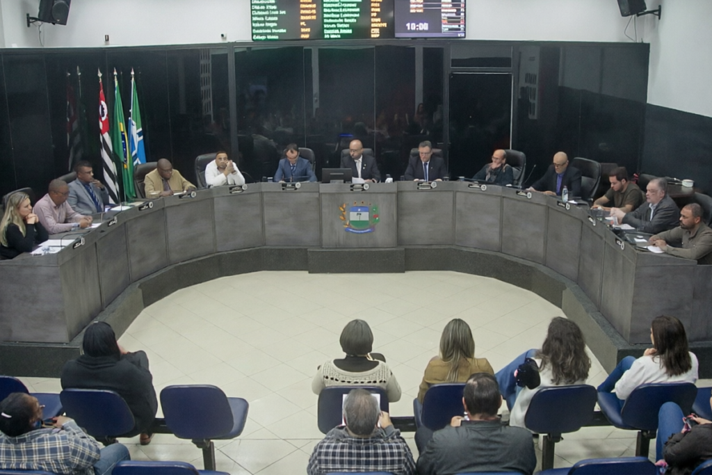 Visão geral do plenário de uma câmara municipal de Jarinu durante uma sessão, onde vereadores estão dispostos em uma grande bancada em formato de semi-círculo cinza. Ao fundo, veem-se bandeiras oficiais e um painel eletrônico de votação, enquanto o público assiste de costas em cadeiras azuis no primeiro plano.