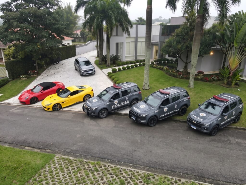 Visão aérea de uma operação policial em uma casa de alto padrão. Alinhados na rua, em frente ao gramado da residência, estão três SUVs cinzas da Polícia Militar (BAEP). No pátio de cascalho da casa, aparecem dois carros esportivos de luxo — um Porsche conversível vermelho e uma Ferrari amarela — além de um SUV cinza ao fundo. A casa ao fundo possui arquitetura moderna, com grandes janelas de vidro e paisagismo com palmeiras.