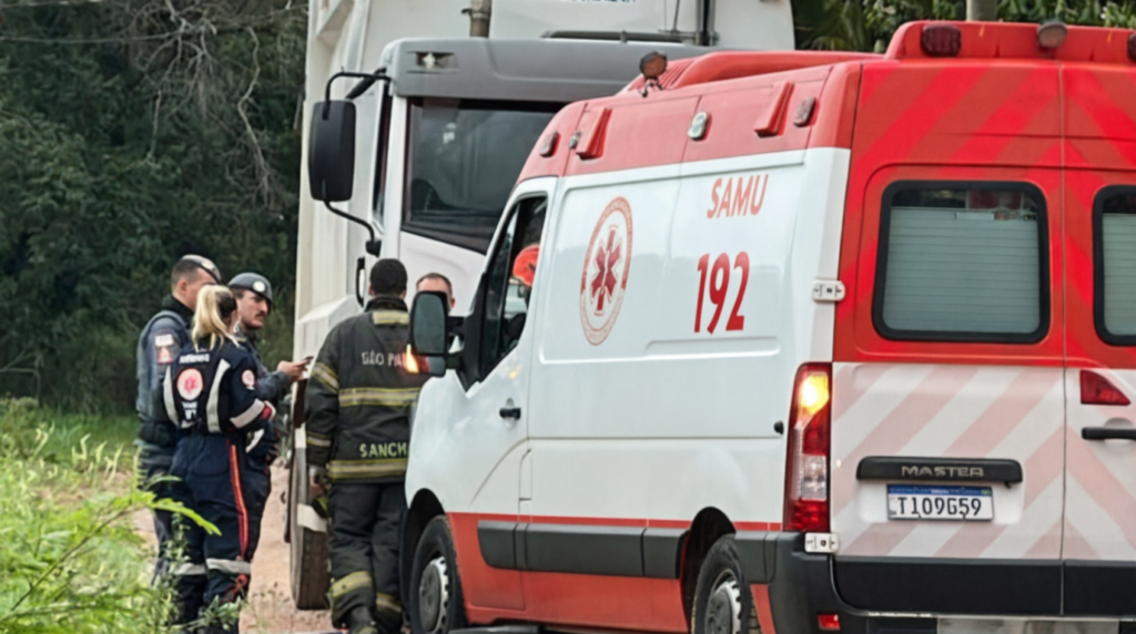Fotografia de atendimento de emergência em via pública. Em primeiro plano, vê-se a parte traseira e lateral de uma ambulância branca e vermelha do SAMU 192. Ao fundo, estão estacionados um caminhão e socorristas, além de policiais militares e bombeiros de São Paulo conversando no local da ocorrência.
