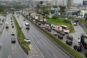 Vista aérea de uma rodovia movimentada em um dia chuvoso. O tráfego no sentido superior está com congestionamento intenso, com carros, ônibus e caminhões em fila, enquanto as faixas no sentido oposto têm fluxo livre. Ao fundo, veem-se prédios, estabelecimentos comerciais como uma concessionária da Iveco e vegetação sob céu nublado.