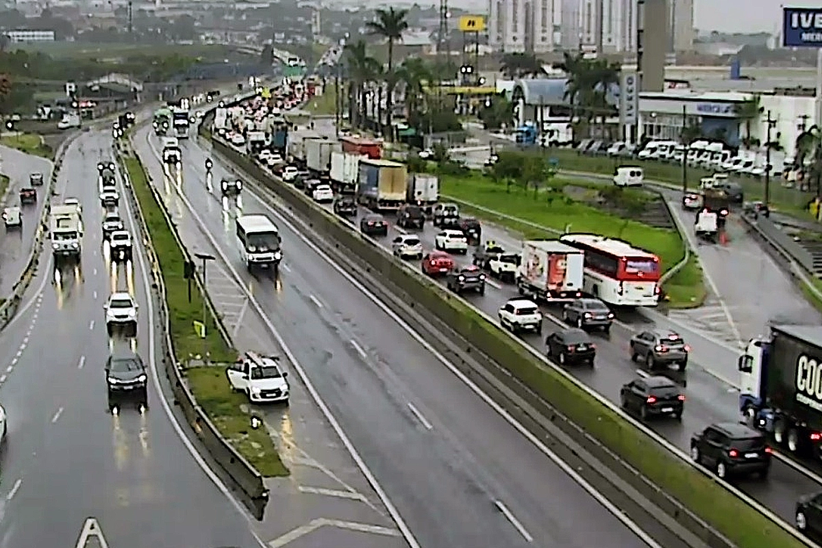 Vista aérea de uma rodovia movimentada em um dia chuvoso. O tráfego no sentido superior está com congestionamento intenso, com carros, ônibus e caminhões em fila, enquanto as faixas no sentido oposto têm fluxo livre. Ao fundo, veem-se prédios, estabelecimentos comerciais como uma concessionária da Iveco e vegetação sob céu nublado.