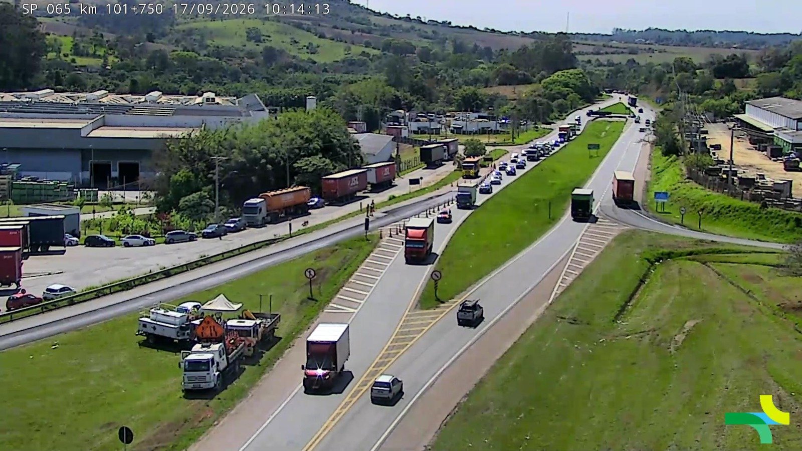 Imagem de câmera de monitoramento rodoviário mostrando o tráfego na rodovia SP-065, no km 101+750. A cena exibe trechos com trânsito intenso e retenção de veículos, além de caminhões e carros em movimento em uma área cercada por vegetação e galpões industriais.