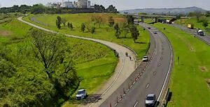 Imagem panorâmica capturada por câmera de monitoramento viário mostrando uma alça de acesso curvada que conecta a uma rodovia movimentada. No acostamento e ao longo da pista, veem-se cones de sinalização delimitando a via, um carro branco parado na vegetação ao lado da curva e outros veículos trafegando pela pista principal. Ao fundo, observam-se áreas verdes, viadutos e construções em um dia ensolarado.