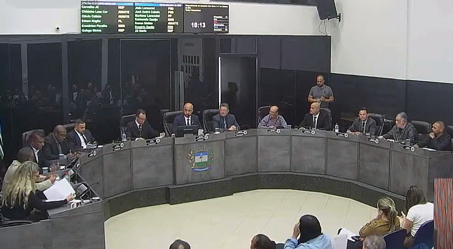 Vereadores reunidos no plenário da Câmara Municipal durante sessão legislativa, dispostos em uma bancada semi-circular com o brasão do município ao centro e painel eletrônico ao fundo.