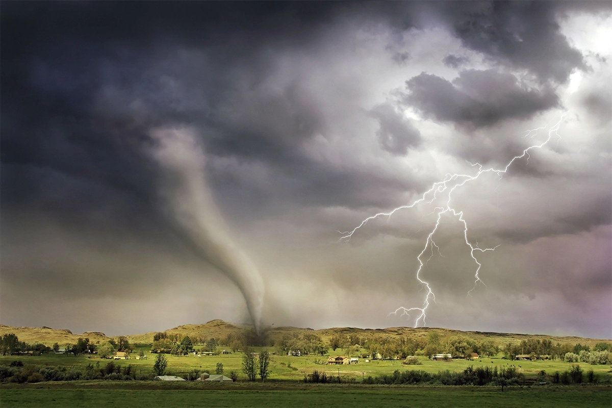 ornado em formato de funil descendo de céus tempestuosos e atingindo uma área rural, acompanhado por um grande raio cortando as nuvens escuras ao lado.